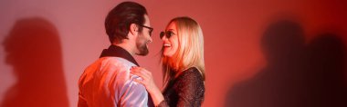 Stylish woman in sunglasses hugging boyfriend on red background, banner 