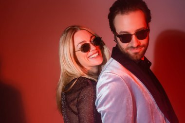 Smiling blonde woman standing near stylish boyfriend during party on red background