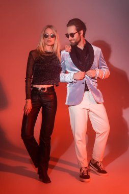 Full length of stylish man buttoning jacket near girlfriend in sunglasses during party on red background 