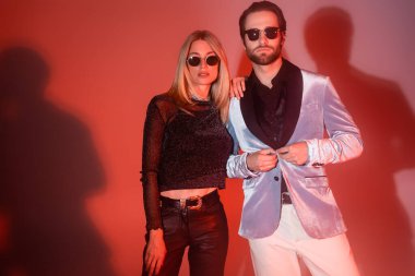 Stylish woman in sunglasses hugging boyfriend in jacket on red background 