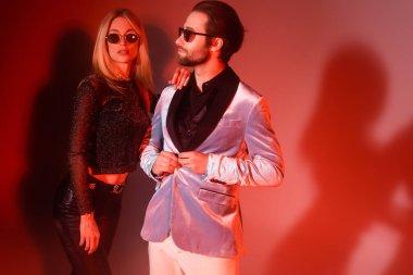 Trendy woman in sunglasses hugging boyfriend in jacket during party on red background 