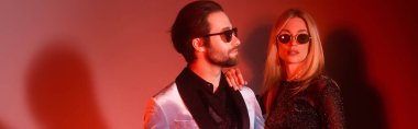 Blonde woman in sunglasses hugging boyfriend in jacket during party on red background, banner 