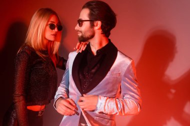 Stylish woman hugging trendy boyfriend in sunglasses during party on red background 