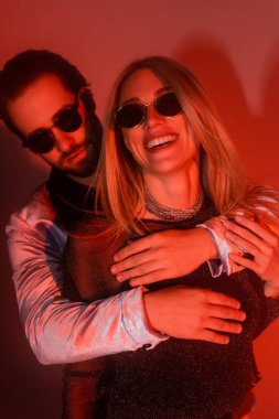 Man in sunglasses embracing cheerful girlfriend during party on red background 