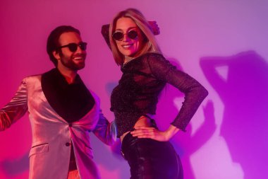 Cheerful blonde woman in sunglasses dancing near boyfriend during party on purple background 