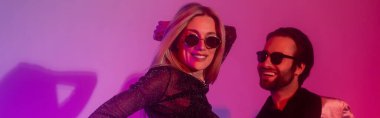 Smiling woman in sunglasses dancing with boyfriend during party on purple background, banner  
