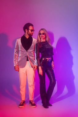 Trendy couple in sunglasses standing on purple background during party 