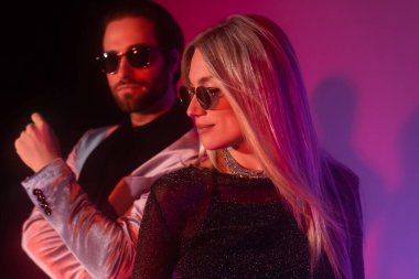 Stylish woman in sunglasses standing near blurred boyfriend during party on purple background 