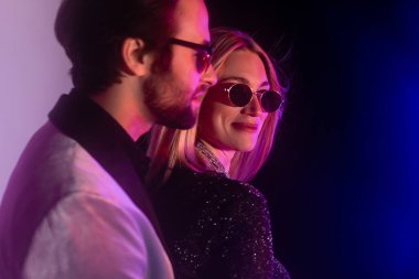 Smiling woman in sunglasses standing near blurred boyfriend on purple and blue background 