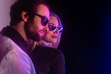 Stylish man in sunglasses looking away near blurred girlfriend during party on purple and black background