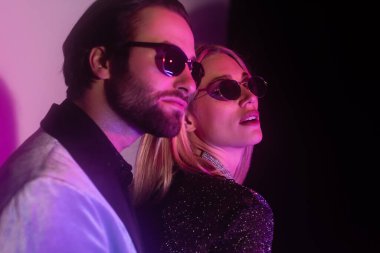 Stylish woman in sunglasses standing near blurred boyfriend on purple and black background 