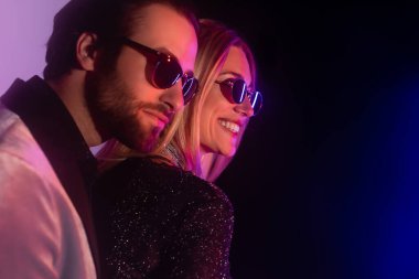 Cheerful blonde woman in sunglasses standing near bearded boyfriend on purple and black background 