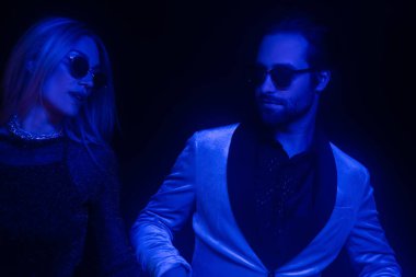 Stylish couple in sunglasses standing isolated on black with blue lighting 