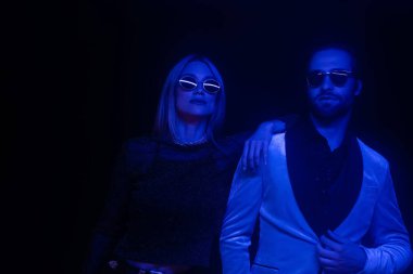 Fashionable couple in sunglasses standing isolated on black with blue lighting 