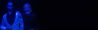 Trendy couple in sunglasses standing during party isolated on black with blue lighting, banner 