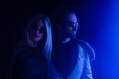 Blonde woman in sunglasses hugging stylish boyfriend on black with blue lighting 