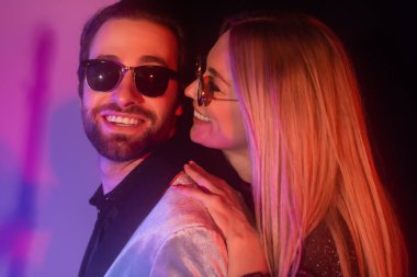 Cheerful blonde woman in sunglasses hugging boyfriend in jacket on purple background 