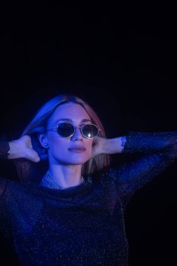 Stylish woman in sunglasses touching hair isolated on black with blue lighting 