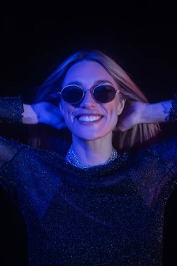Smiling woman in sunglasses touching hair isolated on black with blue lighting 