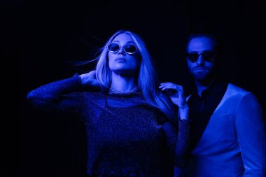 Trendy blonde woman standing near boyfriend in sunglasses isolated on black with blue lighting 
