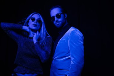 Stylish woman touching neck near boyfriend in sunglasses isolated on black with blue lighting 