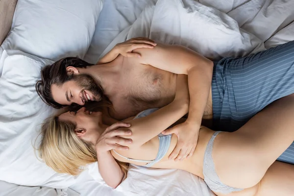 top view of smiling man in pajama pants embracing seductive woman in sexy lingerie on bed