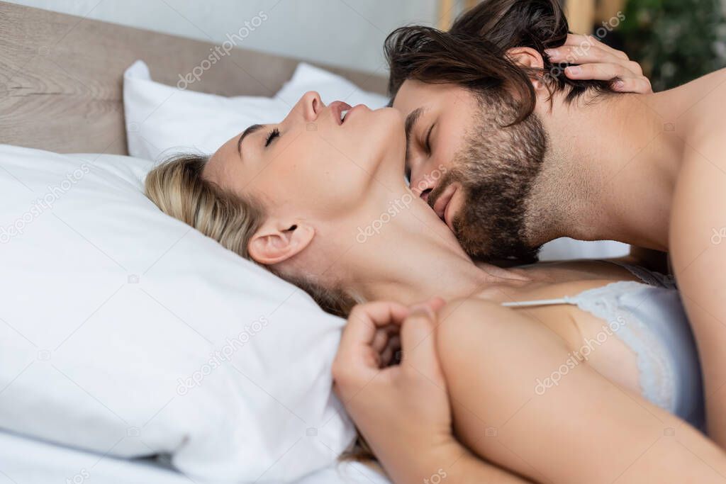 hot woman lying on bed with closed eyes near man kissing her neck 