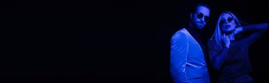 Fashionable man in sunglasses standing near girlfriend in sunglasses isolated on black with blue lighting, banner 