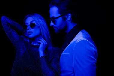 Man in jacket and sunglasses standing near stylish girlfriend isolated on black with blue lighting 