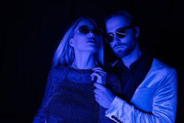 Young man in sunglasses touching hand of blonde girlfriend during party isolated on black with blue lighting 
