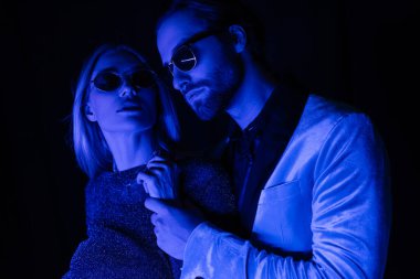 Stylish young man touching hand of girlfriend in sunglasses isolated on black with blue lighting 