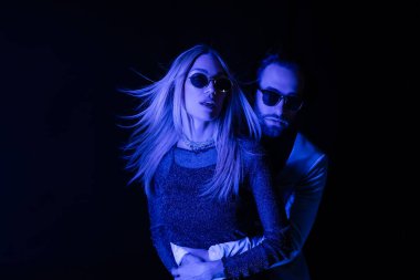 Man in sunglasses hugging blonde girlfriend during party isolated on black with blue lighting 