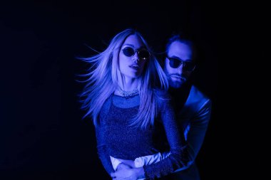 Man in sunglasses hugging blonde girlfriend during party isolated on black with blue lighting 