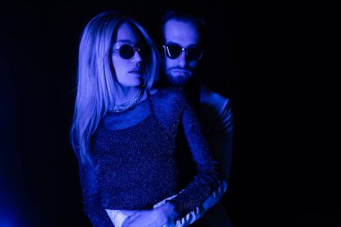Young man hugging stylish woman during party isolated on black with blue lighting 