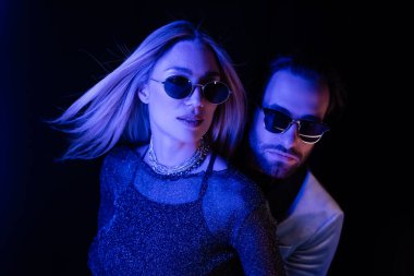 Stylish couple in sunglasses standing isolated on black with blue lighting during party 