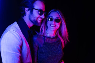 Smiling blonde woman standing near boyfriend in sunglasses isolated on black with colorful lighting 