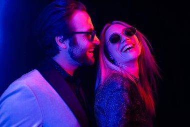 Fashionable man looking at girlfriend in sunglasses isolated on black with colorful lighting 