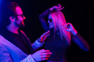 Stylish coupe in sunglasses dancing isolated on black with colorful lighting 
