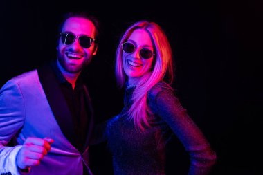 Cheerful blonde woman in sunglasses standing near boyfriend in jacket isolated on black with colorful lighting 