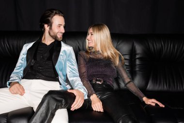 Cheerful stylish couple looking at each other while sitting on leather couch isolated on black 