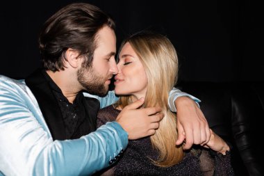 Side view of stylish couple kissing on leather couch during party isolated on black 