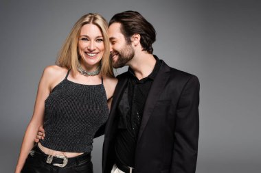 Cheerful man in jacket hugging stylish blonde woman isolated on grey 