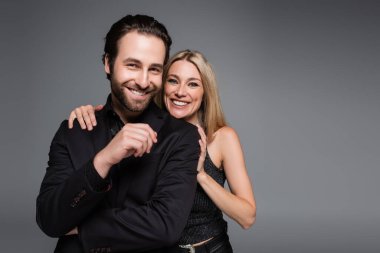 Blonde woman hugging smiling boyfriend in jacket isolated on grey