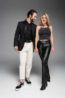 full length of stylish man looking at blonde girlfriend on grey background 
