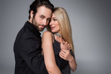 Bearded man in jacket hugging blonde girlfriend isolated on grey 