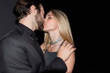Trendy woman kissing bearded boyfriend on black background 