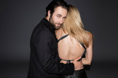 Young man in jacket hugging blonde girlfriend and looking at camera isolated on grey 