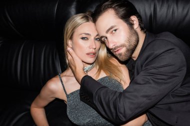 Young man in jacket touching blonde girlfriend on leather couch 