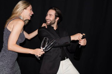 Cheerful woman in dress holding glasses while boyfriend opening champagne isolated on black 