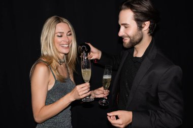 Bearded man pouring champagne near girlfriend in dress isolated on black 